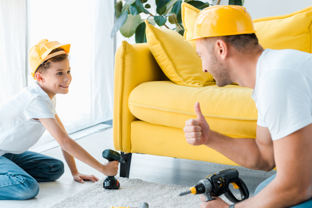 selective focus of happy kid in safety helmet holding hammer drill and looking at father showing thumb up  - Photo, Image