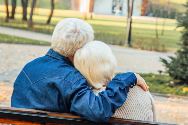 Happy elderly man and woman sitting on a bench in autumn day. Relaxed senior couple sitting on a park bench. Grandfather gently kisses grandmother on the forehead. - Foto, afbeelding