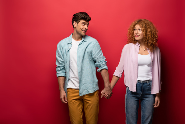 beautiful happy couple in casual clothes holding hands isolated on red - Photo, Image
