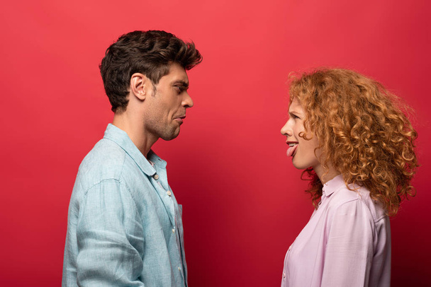 beautiful couple looking at each other and making grimaces, isolated on red - Фото, изображение