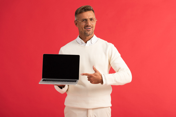 hombre feliz en gafas apuntando con el dedo a la computadora portátil con pantalla en blanco aislado en rojo
  - Foto, imagen