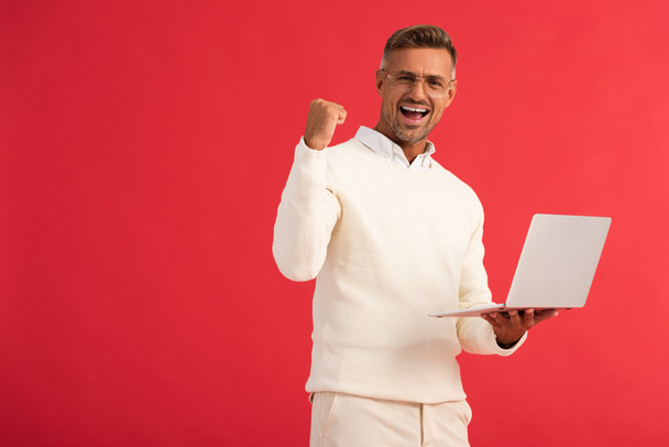 ενθουσιασμένος άνθρωπος σε γυαλιά κρατώντας φορητό υπολογιστή απομονώνονται σε κόκκινο  - Φωτογραφία, εικόνα