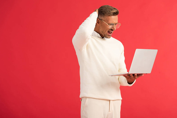 uomo scontento in occhiali che tiene il computer portatile isolato su rosso
  - Foto, immagini