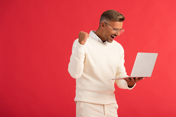 excited and stylish man in glasses holding laptop isolated on red  - Фото, изображение