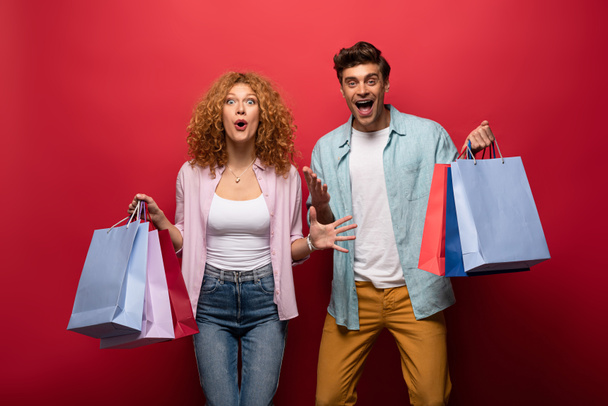 excited beautiful couple holding shopping bags, isolated on red - Fotó, kép