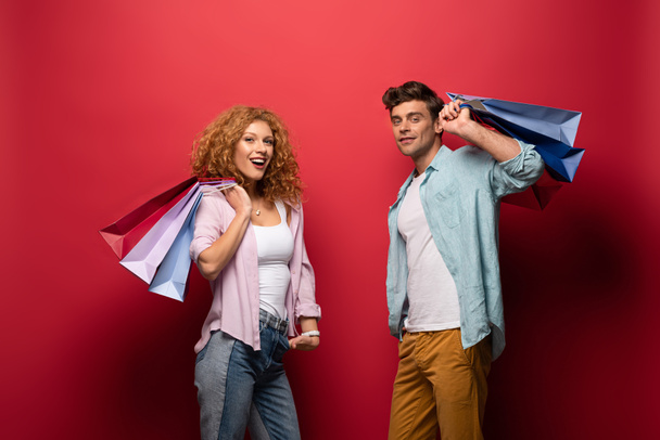 cheerful couple holding shopping bags, isolated on red - Foto, Imagem