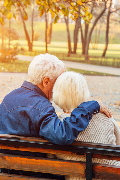 Happy elderly man and woman sitting on a bench in autumn day. Relaxed senior couple sitting on a park bench. Grandfather gently kisses grandmother on the forehead. - Fotó, kép