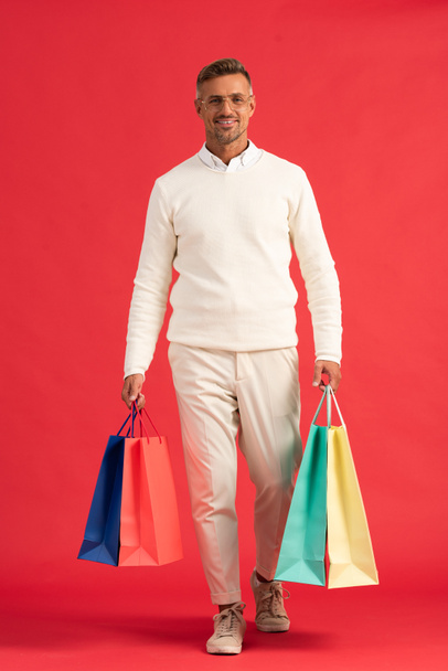 cheerful man in glasses holding colorful shopping bags on red  - Foto, Imagen