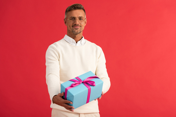 positive man in glasses holding gift box isolated on red  - Фото, изображение