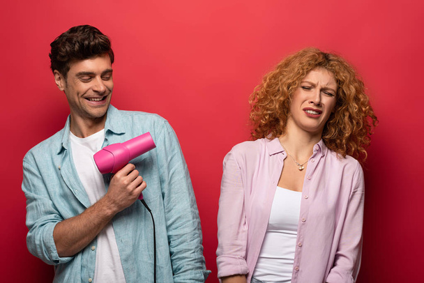 beautiful couple having fun with hair dryer, isolated on red - Φωτογραφία, εικόνα