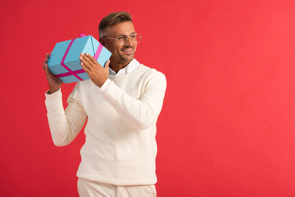 happy man in glasses holding gift box isolated on red  - Foto, Imagen
