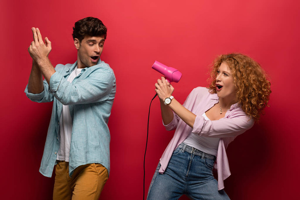 young couple having fun with hair dryer and hand gun, on red - Photo, Image