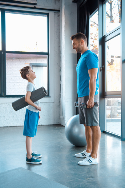 Seitenansicht eines Kindes, das Vater beim Stehen mit Fitnessmatte im Fitnessstudio ansieht  - Foto, Bild