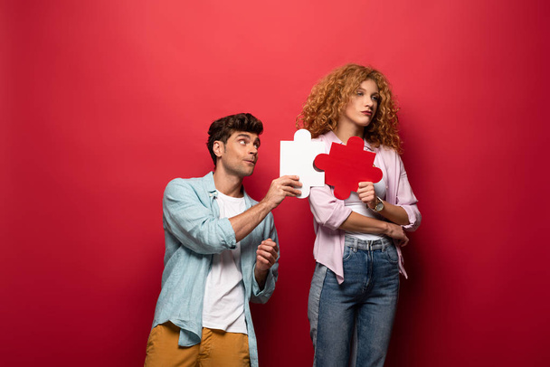 zdenerwowana kobieta i mężczyzna trzymając kawałki układanki, izolowane na czerwono - Zdjęcie, obraz