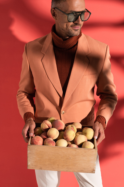 happy man in sunglasses holding box with sweet peaches on red with shadows - Photo, Image