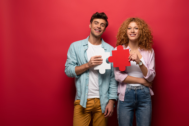 cheerful couple holding puzzle pieces, isolated on red - Photo, Image
