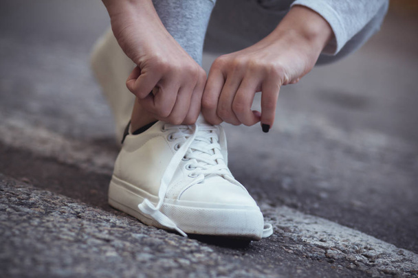 girl squatted down to tie shoelaces on white sneakers on asphalt road, autumn sport concept outdoors - Zdjęcie, obraz