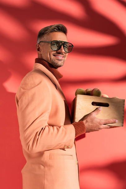 happy man in blazer and sunglasses holding box with sweet peaches on red with shadows - Fotografie, Obrázek