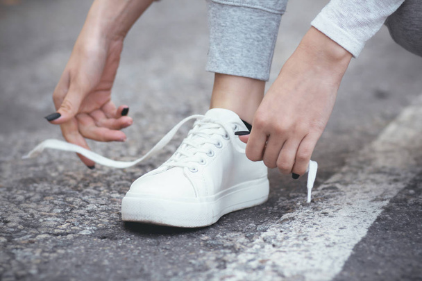 girl squatted down to tie shoelaces on white sneakers on asphalt road, autumn sport concept outdoors - Photo, Image