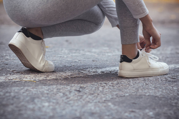 girl squatted down to tie shoelaces on white sneakers on asphalt road, autumn sport concept outdoors - Φωτογραφία, εικόνα