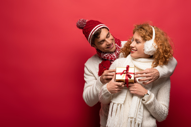 ευτυχισμένο ζευγάρι σε χειμωνιάτικο ντύσιμο κρατώντας χριστουγεννιάτικο δώρο απομονωμένο στο κόκκινο - Φωτογραφία, εικόνα