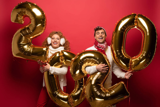 beautiful couple in winter outfit holding new year 2020 golden balloons, on red - Φωτογραφία, εικόνα