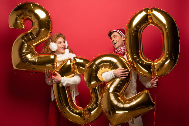 χαμογελαστό ζευγάρι σε χειμερινό ντύσιμο εκμετάλλευση νέο έτος 2020 χρυσά μπαλόνια, σε κόκκινο - Φωτογραφία, εικόνα