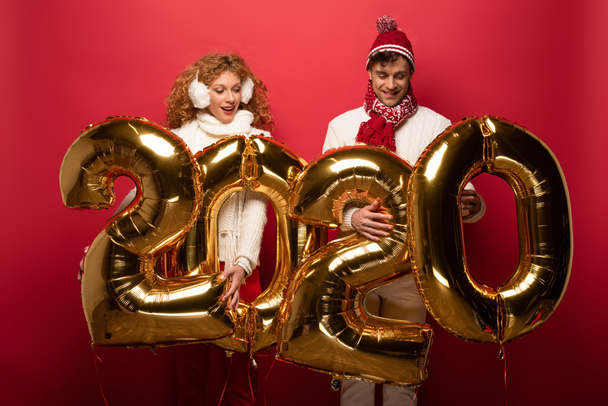 happy couple in winter outfit holding new year 2020 golden balloons, on red - Fotografie, Obrázek
