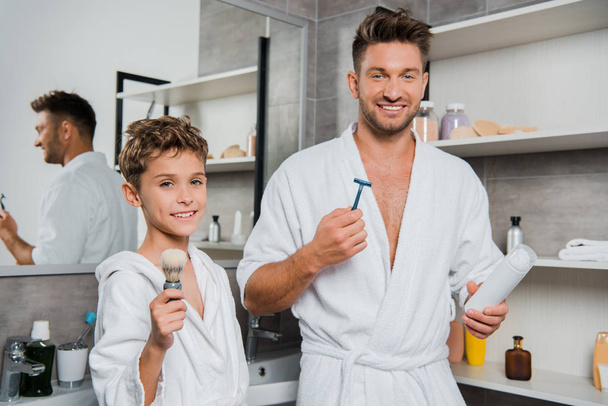 feliz padre sosteniendo la navaja y puede con espuma de afeitar cerca de hijo con cepillo de afeitar
  - Foto, Imagen