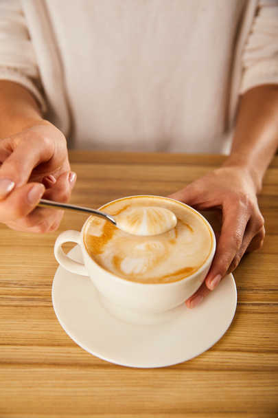 selective focus of woman holding spoon with cappuccino foam near wooden table  - Foto, Imagen
