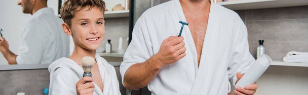 plano panorámico de padre sosteniendo la afeitadora y puede con espuma de afeitar cerca hijo feliz con cepillo de afeitar
  - Foto, Imagen