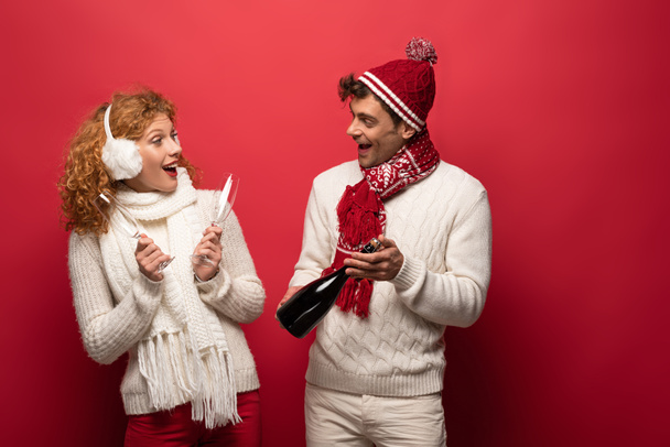 прекрасна щаслива пара в зимовому одязі, що тримає шампанське, щоб відсвяткувати хрещення, на червоному. - Фото, зображення