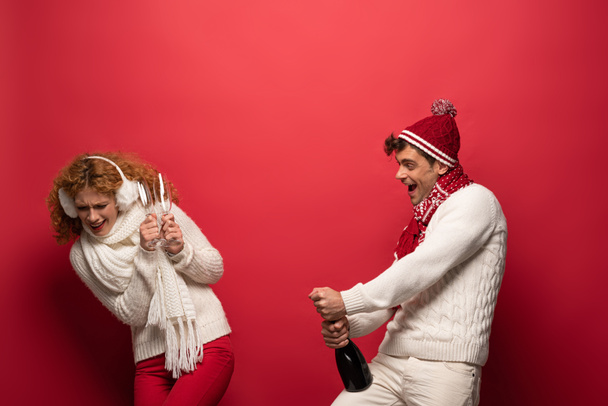 cheerful funny couple in winter outfit holding champagne for celebrating christmas, on red - Foto, afbeelding