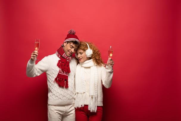beautiful couple in winter outfit hugging and holding champagne glasses, on red - Fotografie, Obrázek