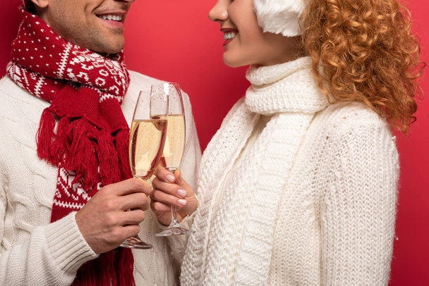 oříznutý pohled na usmívající se pár v zimním oblečení cinkající sklenicemi šampaňského, izolované na červené - Fotografie, Obrázek