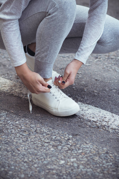 Kız asfalt yolda beyaz spor ayakkabılara ayakkabı bağlamak için çömelmiş, sonbahar sporu kavramı açık havada. - Fotoğraf, Görsel