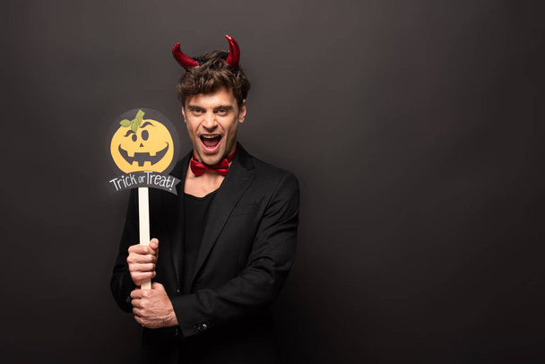 sexy man in devil costume holding pumpkin trick or treat sign on black - Valokuva, kuva