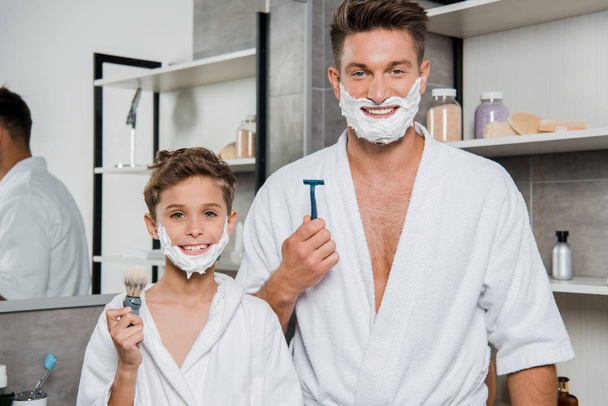 panoramic shot of father and son with shaving foam on faces holding razor and shaving brush  - Foto, afbeelding