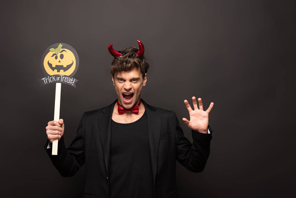 handsome sexy man in halloween devil costume holding pumpkin trick or treat sign on black - Photo, Image