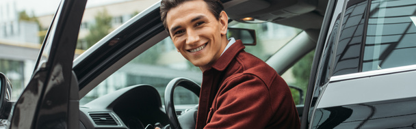 Taxi chauffeur zittend in auto met open deur en glimlachend op camera, panoramische opname - Foto, afbeelding