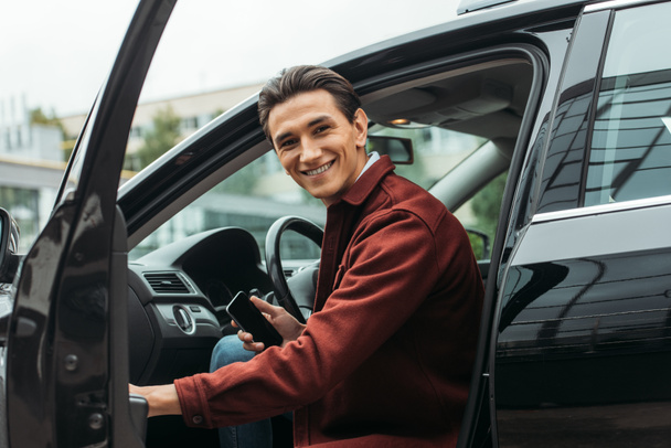 Hymyilevä taksinkuljettaja istuu autossa, jossa on avoin ovi ja jolla on älypuhelin
 - Valokuva, kuva