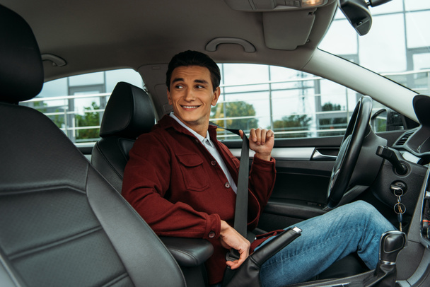 Uomo sorridente che tiene la cintura di sicurezza in auto e distoglie lo sguardo
 - Foto, immagini