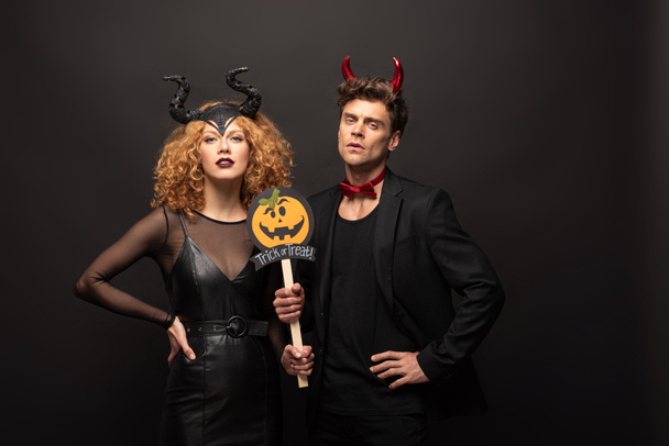 beautiful couple posing in halloween costumes with pumpkin trick or treat sign on black - Photo, Image