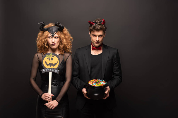 thoughtful couple in halloween costumes holding pot with candies and pumpkin trick or treat sign on black - Фото, изображение