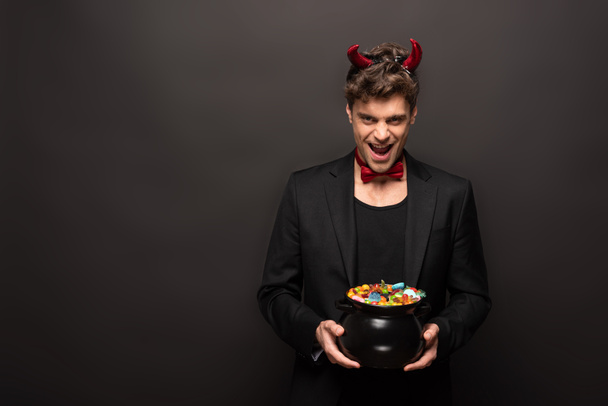 guapo riendo hombre en traje de diablo sosteniendo olla con caramelos en negro
 - Foto, Imagen