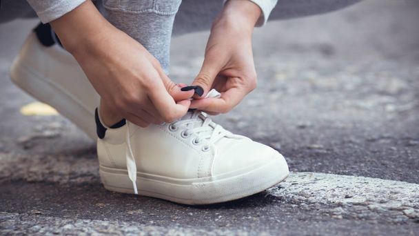 girl squatted down to tie shoelaces on white sneakers on asphalt road, autumn sport concept outdoors - 写真・画像