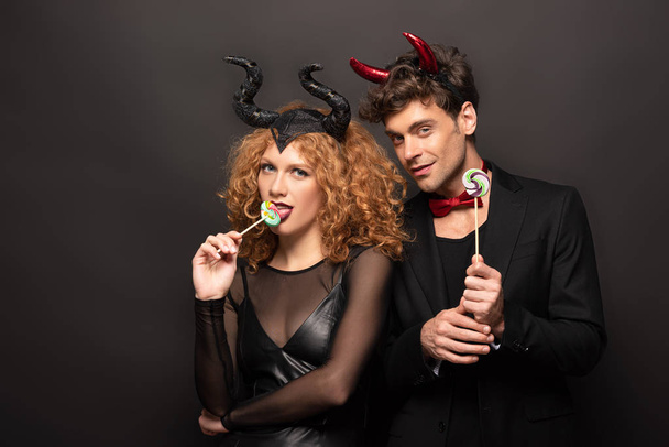 sexy couple in halloween costumes eating lollipops on black - Foto, imagen