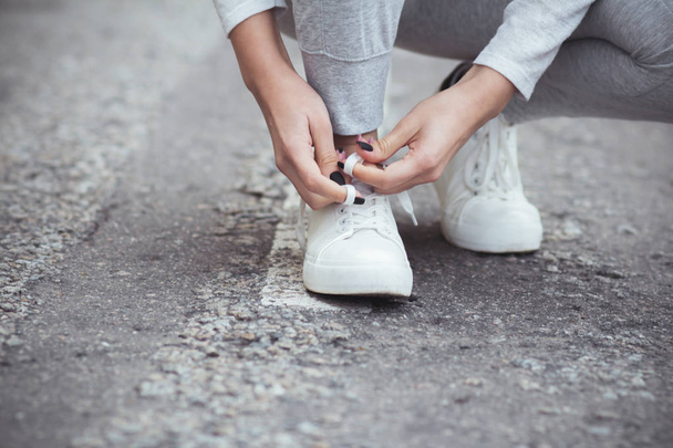 girl squatted down to tie shoelaces on white sneakers on asphalt road, autumn sport concept outdoors - Zdjęcie, obraz
