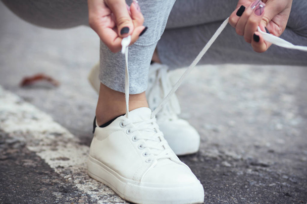 girl squatted down to tie shoelaces on white sneakers on asphalt road, autumn sport concept outdoors - Foto, imagen