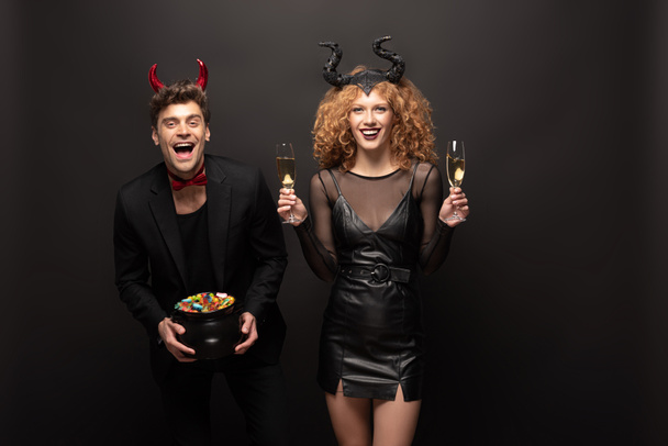 happy couple posing in halloween costumes with glasses of champagne and pot of candies on black - Φωτογραφία, εικόνα